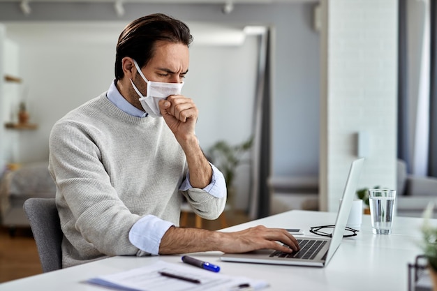 Joven empresario tosiendo mientras usa mascarilla protectora y trabaja en una computadora en casa