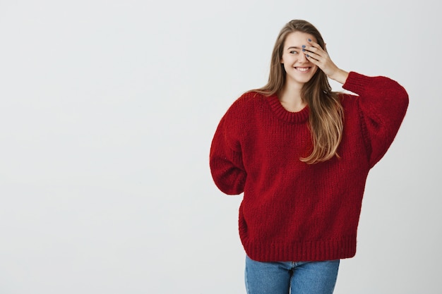 Joven y enamorado. Adorable mujer guapa con suéter rojo suelto que esconde la mano detrás y se cubre un ojo mientras mira a la izquierda y sonríe juguetonamente, de pie