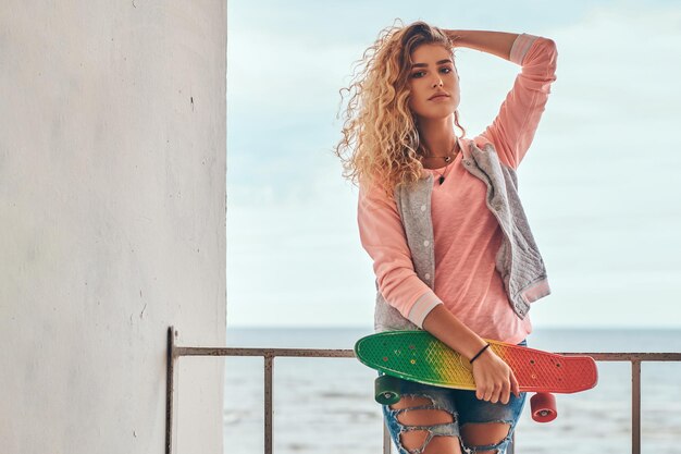 Una joven encantadora con cabello rubio vestido con una chaqueta rosa sostiene un monopatín posando cerca de una barandilla contra la costa del mar.