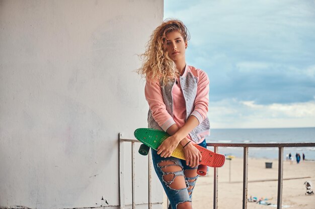 Una joven encantadora con cabello rubio vestido con una chaqueta rosa sostiene un monopatín posando cerca de una barandilla contra la costa del mar.