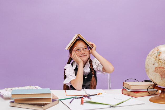 Una joven y encantadora colegiala con coletas y pecas en gafas transparentes mirando hacia otro lado y sosteniendo un libro en la cabeza sobre un fondo aislado