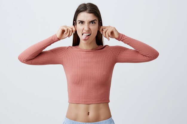 Joven encantadora divertida alegre niña caucásica con largo cabello castaño en camisa rosa y pantalones cortos azules tirando de las orejas con las manos, mostrando la lengua, haciendo una expresión de cara divertida para que la hermanita deje de llorar.