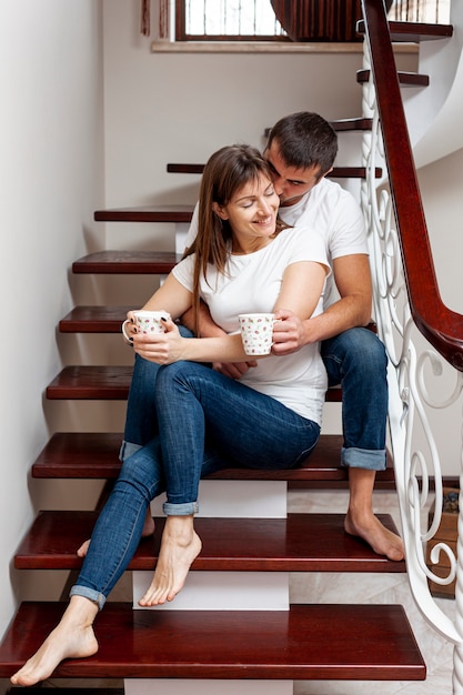 Joven encantadora pareja sosteniendo tazas de café y sentado en las escaleras
