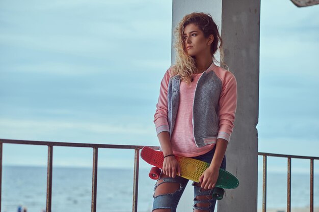 Una joven encantadora con el pelo rubio vestido con una chaqueta rosa sostiene un monopatín posando cerca de una barandilla contra la costa del mar, mirando hacia otro lado.