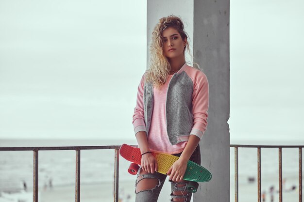 Una joven encantadora con el pelo rubio vestido con una chaqueta rosa sostiene un monopatín posando cerca de una barandilla contra el fondo de la costa del mar en blanco y negro.