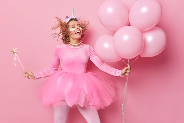 Una joven enérgica y optimista sostiene un montón de globos inflados, tiene una expresión alegre, usa un vestido festivo plisado, se somete a procedimientos de belleza y disfruta de la fiesta aislada en un fondo rosa