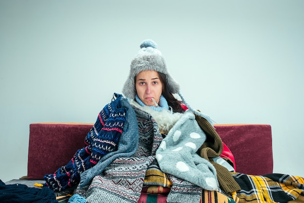 Foto gratuita la joven enferma con chimenea sentada en el sofá en casa o estudio cubierto con ropa de abrigo tejida. enfermedad, influenza, concepto de dolor. relajación en casa. conceptos sanitarios.