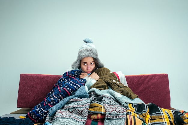 Foto gratuita la joven enferma con chimenea sentada en el sofá en casa o estudio cubierto con ropa de abrigo tejida. enfermedad, influenza, concepto de dolor. relajación en casa. conceptos sanitarios.