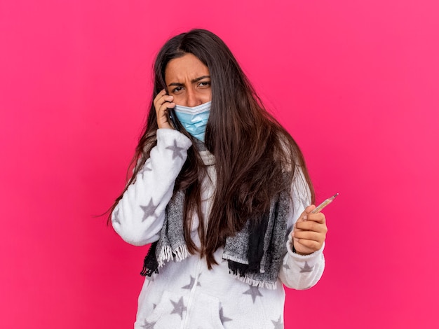 Joven enferma molesta mirando a cámara con máscara médica con bufanda habla por teléfono y sosteniendo termómetro aislado sobre fondo rosa
