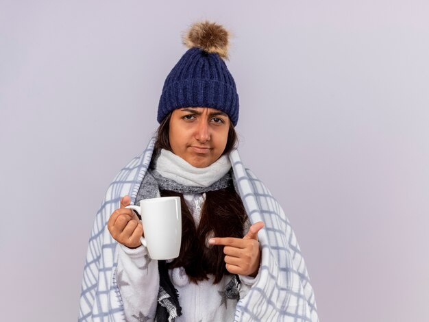 Joven enferma molesta vistiendo gorro de invierno con bufanda envuelta en plaid sosteniendo y apunta a una taza de té