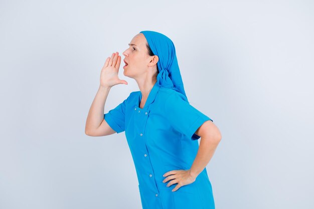 Joven enfermera grita sosteniendo la mano cerca de la boca sobre fondo blanco.