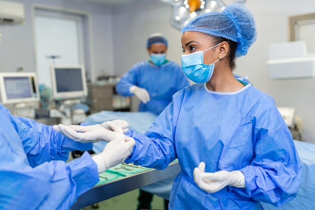 Joven enfermera con máscara protectora y ropa de trabajo ayudando al cirujano con guantes mientras se preparan para la operación