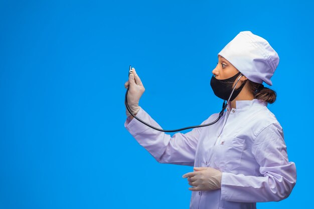 Joven enfermera en mascarilla negra comprueba al paciente con estetoscopio sobre fondo azul.