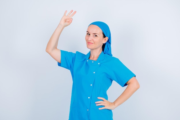 La joven enfermera posa ante la cámara mostrando un gesto correcto y poniendo la otra mano en la cintura sobre fondo blanco.