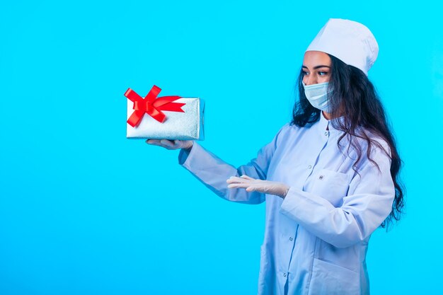 Joven enfermera en uniforme aislado sosteniendo una caja de regalo con cinta roja y presentándola.