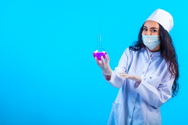 Joven enfermera en uniforme aislado sosteniendo un matraz de prueba sobre superficie azul