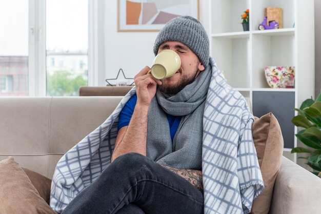 Joven enfermo con bufanda y gorro de invierno sentado en el sofá en la sala de estar envuelto en una manta bebiendo una taza de té con los ojos cerrados