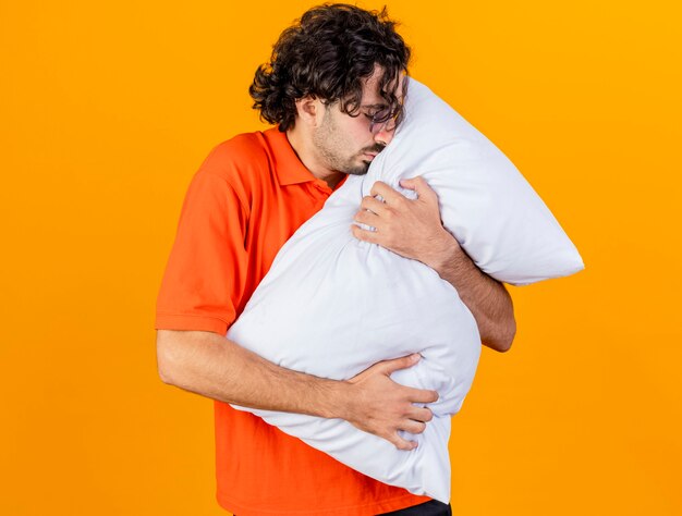 Joven enfermo caucásico con gafas abrazando la almohada tocando la cara con los ojos cerrados aislado sobre fondo naranja con espacio de copia