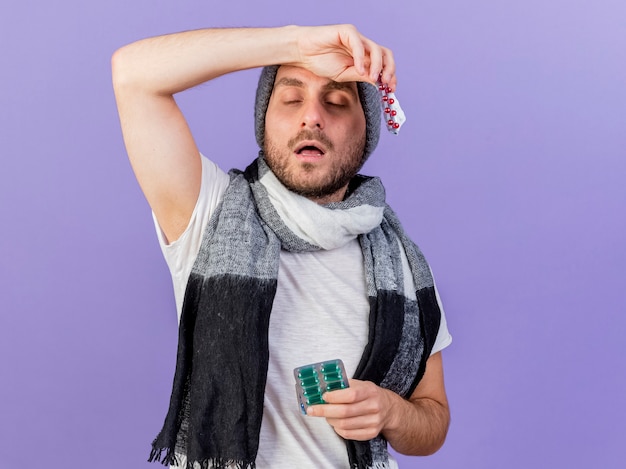 Joven enfermo con gorro de invierno con bufanda sosteniendo pastillas y poniendo el brazo en la frente aislado en púrpura