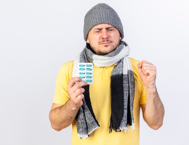 Joven enfermo rubio molesto con gorro y bufanda de invierno sostiene un paquete de píldoras médicas y mantiene el puño aislado en la pared blanca