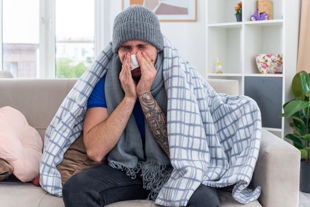 Foto gratuita joven enfermo vestido con bufanda y gorro de invierno envuelto en una manta sentado en el sofá en la sala de estar limpiando la nariz con una servilleta con los ojos cerrados