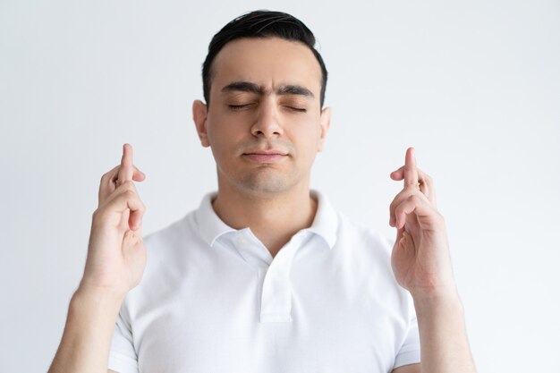 Joven enfocado manteniendo los dedos cruzados y los ojos cerrados. Concepto de deseo