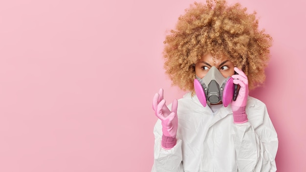 Una joven enojada usa una máscara de gas y un traje de riesgo biológico hace gestos de llamadas telefónicas con molestia preocupada por la contaminación ambiental química aislada sobre el espacio de copia de fondo rosa