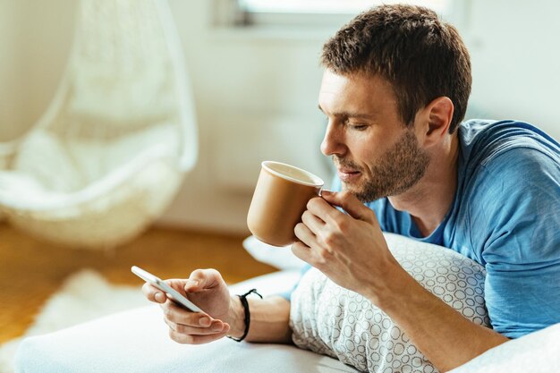 Joven enviando mensajes de texto por teléfono mientras bebe café matutino en la cama