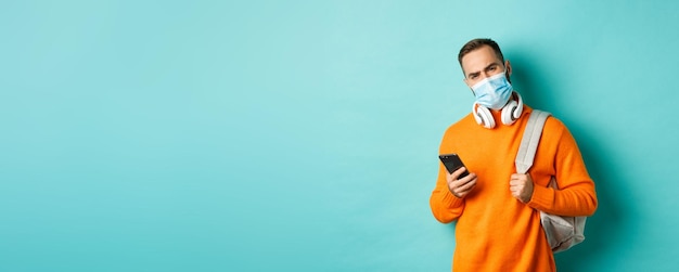 Foto gratuita joven escéptico y decepcionado con mascarilla sosteniendo mochila y teléfono móvil frunciendo el ceño