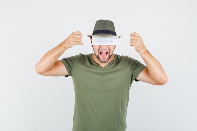 Joven escondiendo los ojos detrás de la máscara médica en camiseta verde y sombrero y mirando divertido