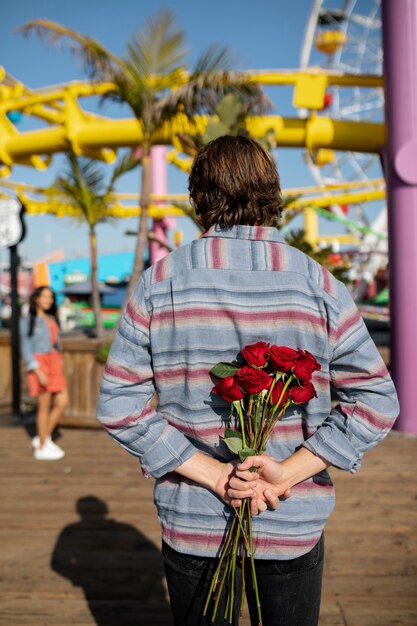 Joven escondiendo un ramo de flores para su novia mientras estaba en una cita en el parque de diversiones