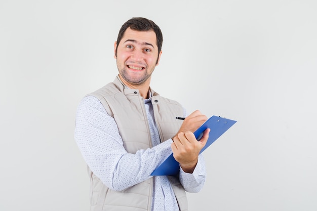 Joven escribiendo algo en el cuaderno con bolígrafo y posando en chaqueta beige y mirando optimista, vista frontal.