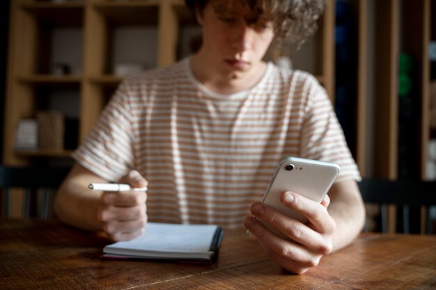 Joven escribiendo algo en un cuaderno mientras mira su teléfono inteligente