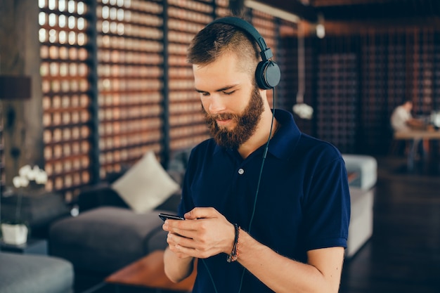 Joven escucha música en auriculares, usando teléfono inteligente, retrato de hipster al aire libre