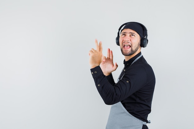 Joven escuchando música con auriculares en camisa, delantal y mirando divertido.