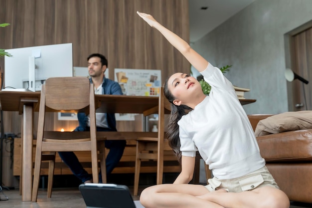 Joven esposa asiática entrena ejercicios de yoga practicando una lección en línea de feomr en la sala de estar con su esposo trabajando detrás del concepto de orden de estado en casa