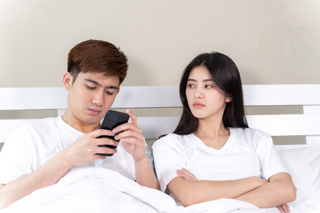 joven esposa sentada en la cama con malestar, su esposo usa el teléfono inteligente en la siesta