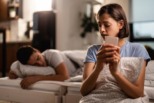 La joven está enviando mensajes de texto en secreto a través de un teléfono inteligente y engañando a su novio que está durmiendo en la cama