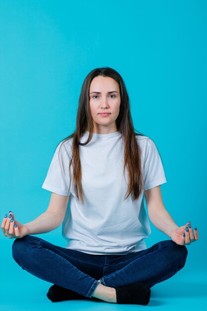 La joven está meditando sentada en el suelo sobre fondo azul.