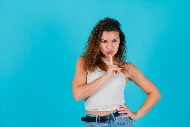 La joven está mirando a la cámara sosteniendo el dedo índice en los labios sobre fondo azul.