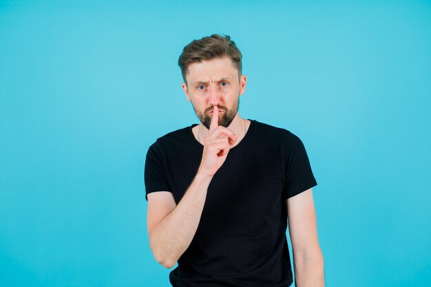 El joven está mostrando un gesto de silencio sosteniendo el dedo índice en los labios sobre fondo azul.