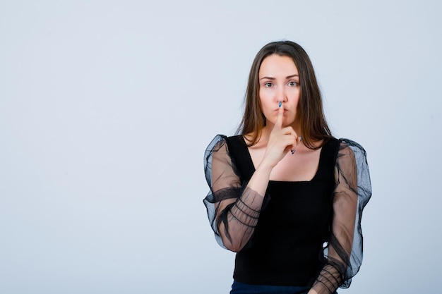 La joven está mostrando un gesto de silencio sosteniendo el dedo índice en los labios sobre fondo blanco.