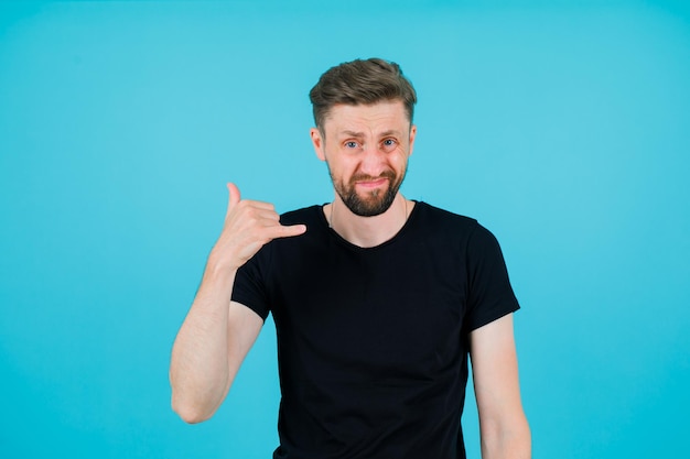 El joven está mostrando un gesto de teléfono sosteniendo la mano cerca de la oreja en el fondo azul