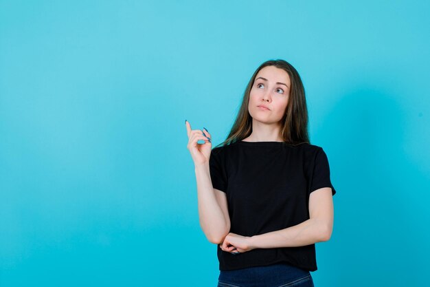 La joven está pensando señalando con el dedo índice sobre fondo azul.