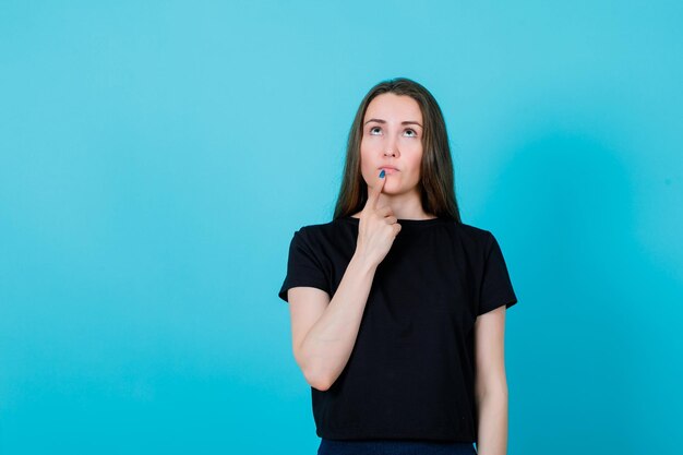 La joven está pensando sosteniendo el dedo índice en los labios sobre fondo azul.