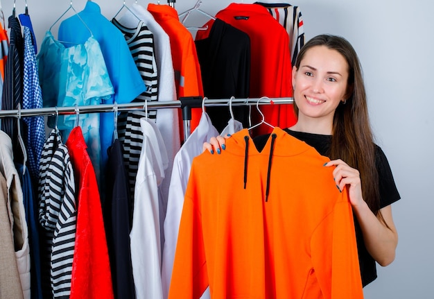 La joven está sonriendo sosteniendo una sudadera en el fondo de la ropa