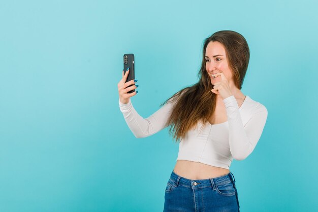 La joven se está tomando una selfie mostrando un gesto de tamaño con la otra mano en el fondo azul