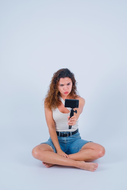 La joven se está tomando selfie con su mini cámara mostrando un mimetismo enojado con fondo blanco