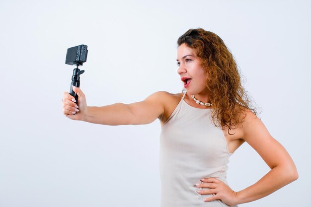 La joven se está tomando selfie con su mini cámara poniendo la mano en la cintura sobre fondo blanco.