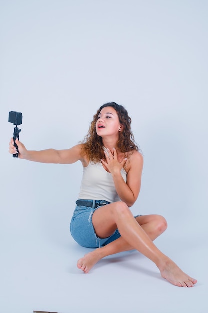 La joven se está tomando una selfie con su mini cámara sosteniendo la mano en el pecho sobre fondo blanco.